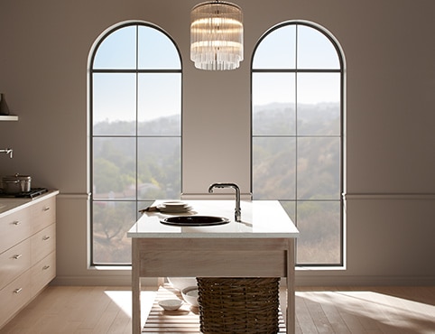 Une cuisine aux murs beiges avec deux grandes fenêtres cintrées et un îlot muni d’un évier.