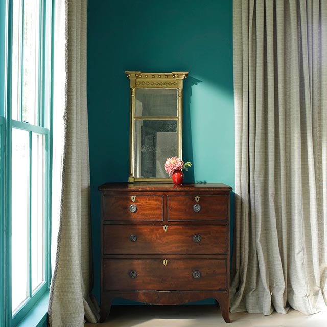 Green-painted bedroom with wooden dresser, gold mirror, and floor-length curtains.