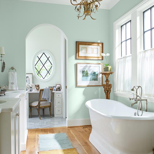 Airy bathroom with pastel-green walls, chandelier, soaking tub, sink and cabinets, wall art, large windows, and a desk and chair.