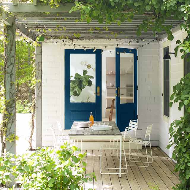 Un porche couvert de lierre avec des portes bleu, des murs blancs, une chaise et une table.