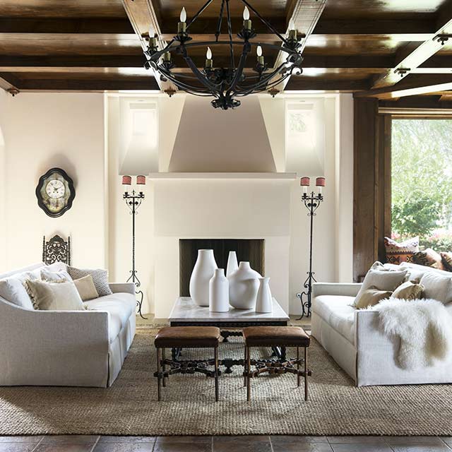 Large open living room with coffered ceiling, chandelier, white seating, leather stools, candelabras, and large windows.