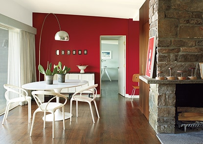 A white midcentury modern dining room with a white round table and chairs, stone fireplace, and a vibrant red accent wall.
