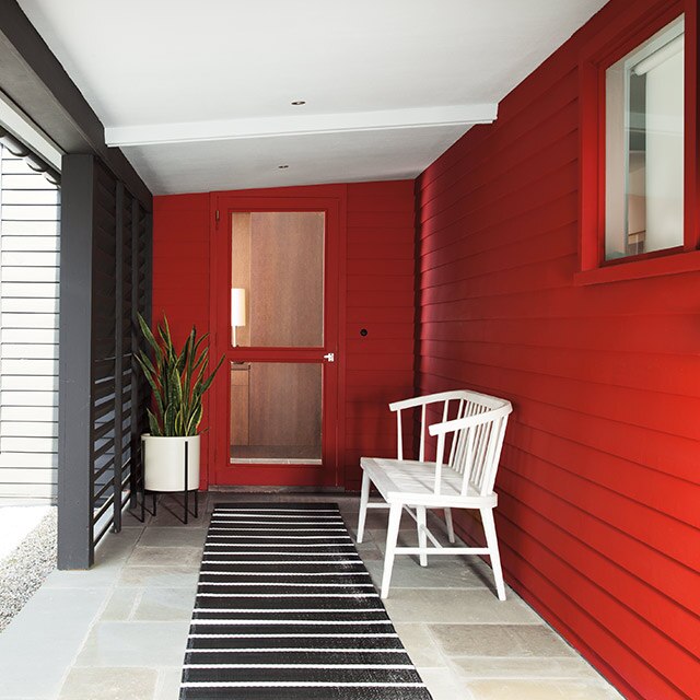 A front porch with a black and white rug and white bench on a house with vibrant red siding and a matching red door.