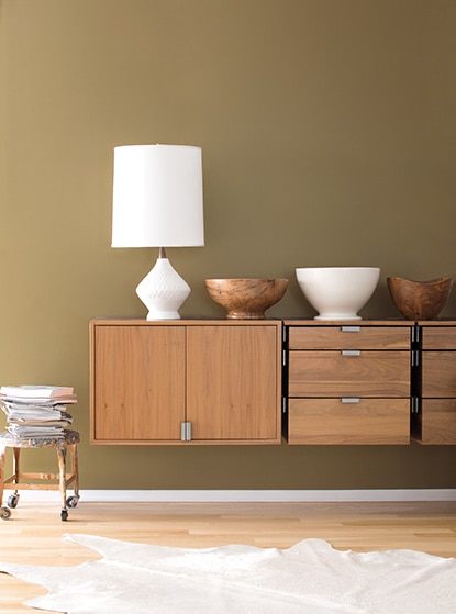 A dark tan wall with three bowls and a white lamp on top of a floating wood cabinet.