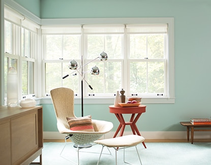 Un coin-lecture aux murs bleu-vert arborant de grandes fenêtres, un fauteuil, un tabouret et une table d’appoint.