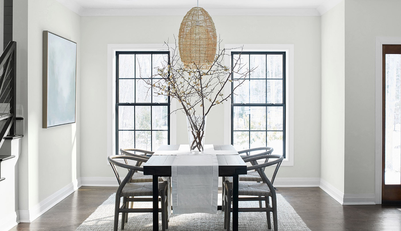 A serene dining room with off-white-painted walls, a light-gray ceiling and trim, black wood table and chairs, and wicker chandelier.