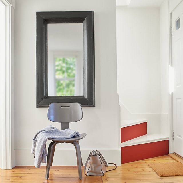 A neat, white-painted entryway and front door, with white stairs, fun red stair risers, a dark gray-framed mirror, gray chair, and blonde wood floor.