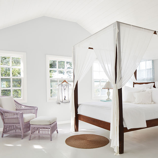 A tranquil, white-painted bedroom with a shiplap vaulted ceiling, a canopy bed and white bedding, purple wicker chair and ottoman, and white wood floor.