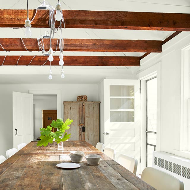 Dining room features a wooden table and white chairs, rustic armoire, wooden counters, and three wooden beams along the ceiling. 