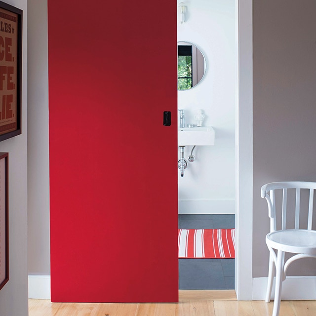 Une chaise d’appoint blanche dans un couloir aux murs gris et une porte de grange rouge qui donne sur une salle de bains arborant une carpette rayée.