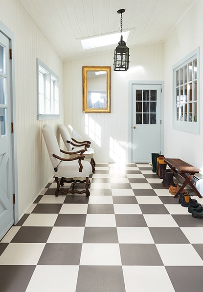 Un couloir arborant un plancher peint en damier gris et blancs, des murs et un plafond blancs, des portes et des moulures bleues, un banc et des chaises.