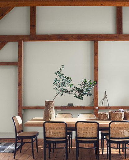 A dining room in neutral hues, with wooden beams, a wooden table, wicker chairs and tabletop baskets.