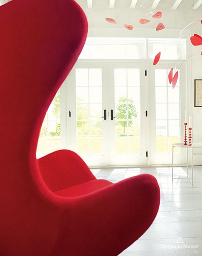 Une pièce lumineuse arborant des murs peints en blanc, un plafond à poutres apparentes, des portes françaises, un fauteuil rouge et un mobile rouge suspendu.