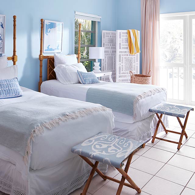 A bedroom with blue painted walls, a white ceiling and trim, twin beds and folding stools in blue and white decor.