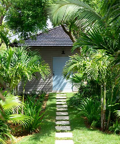 A light gray painted home with a blue door and stone front path, situated among tropical trees and vegetation.