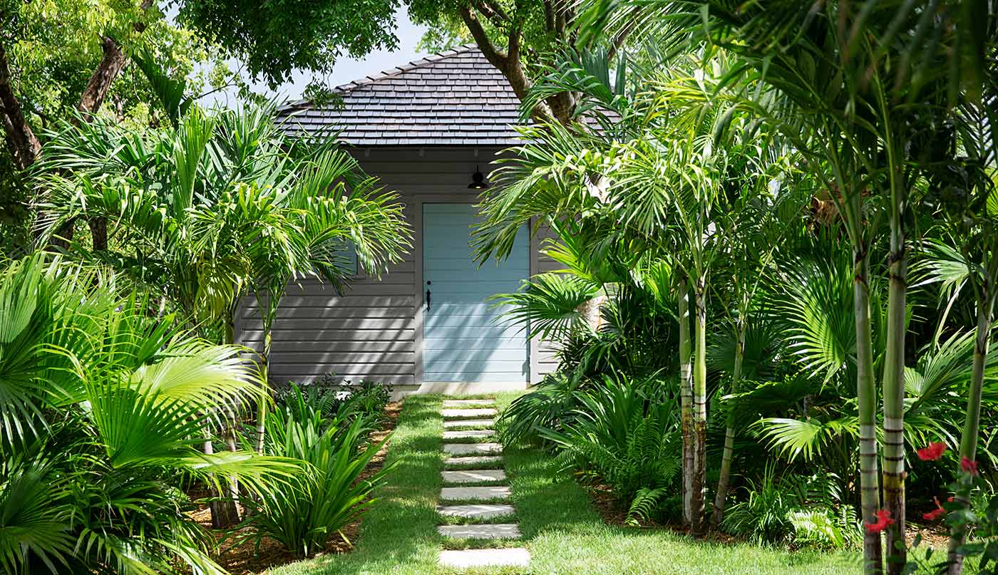 Una casa pintada de gris claro con una puerta azul y un camino frontal de piedra, ubicada entre árboles tropicales y vegetación.