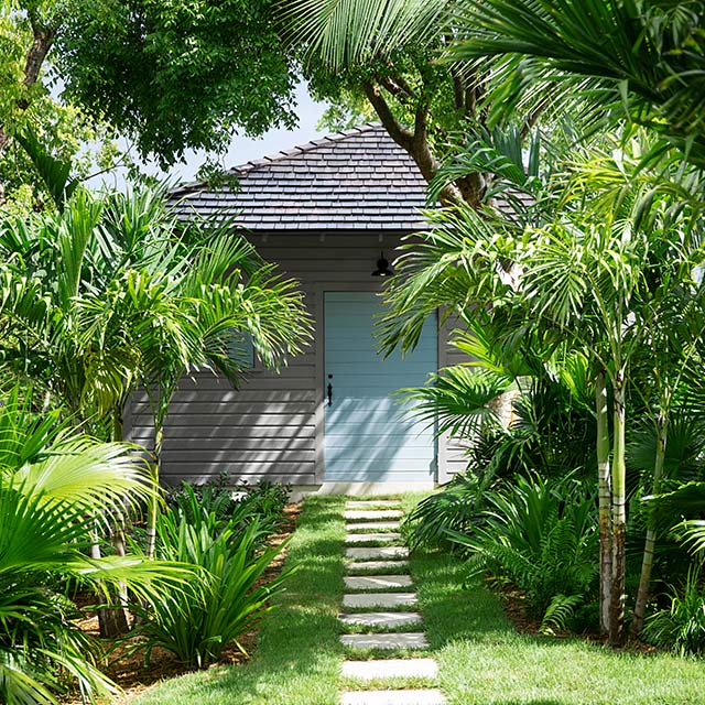 Una casa pintada de gris claro con una puerta azul y un camino frontal de piedra, ubicada entre árboles tropicales y vegetación.