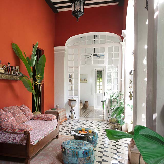 A veranda with orange-painted walls, white posts, trim and doors, a light pink sofa, blue ottomans, and a black-and-white patterned floor.