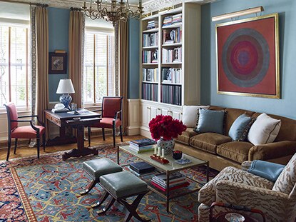 A blue-painted living room with velvet couches, patterned armchair, stools, book shelf, and wall art.