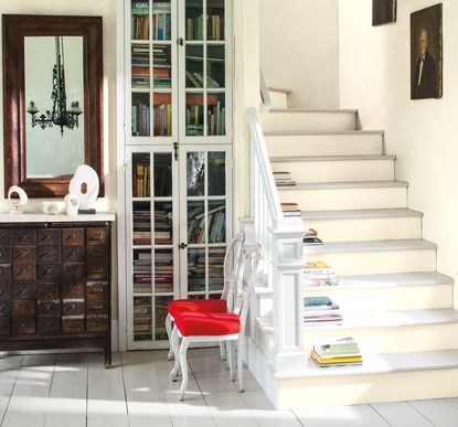 A white-painted living space with a cabinet filled with books, a brown credenza, mirror, and white dining chair with red seat.