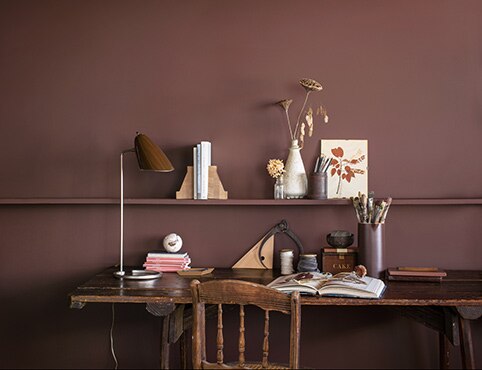 Un bureau aux murs peints en brun arborant une longue tablette en bois, un bureau et une chaise en bois assortis, et diverses décorations.