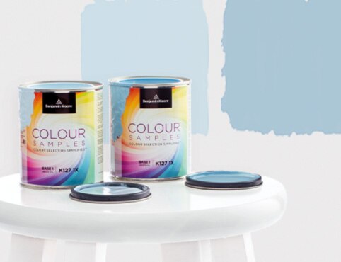 Two open paint colour samples of blue Benjamin Moore paint sitting on a stool, in front of painted squares on a white wall.