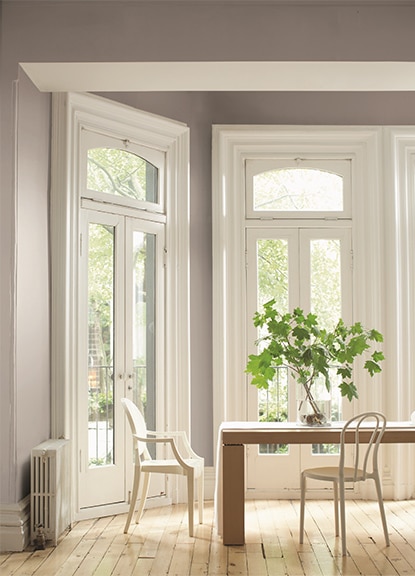 A gray-painted dining room features a wooden plank table, two white chairs, leafy branches in a vase, and French doors.