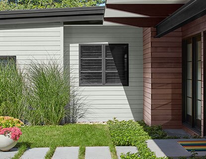 A light, gray-painted modern house with black trim and brown stain, a stone front path, tall grasses, and potted flowers in front.