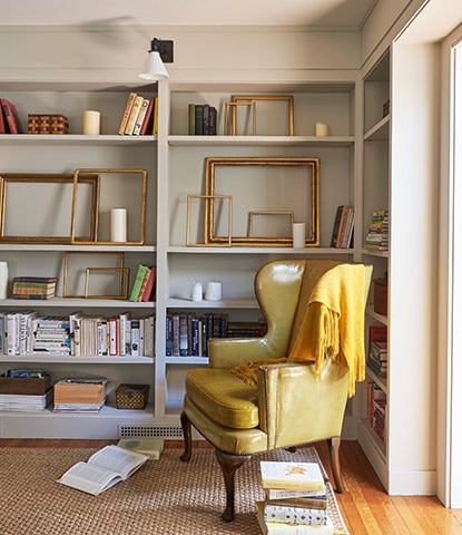 A study with greige-painted walls features alcove bookcases with books and frames, a cozy chair, and golden blanket.