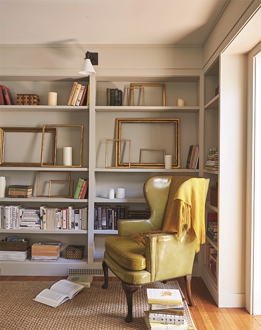 A study with greige-painted walls features alcove bookcases with books and frames, a cozy chair, and golden blanket.