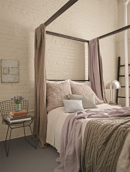 Chambre à coucher avec un mur de briques blanches derrière un lit à baldaquin beige avec des oreillers lilas et une couverture.