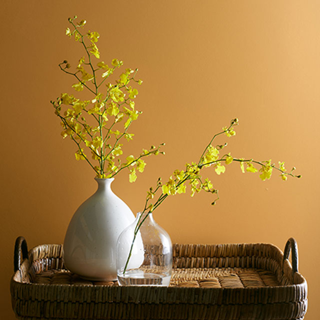 Un vase en céramique et un vase en verre contenant des fleurs jaunes sont posés sur un plateau tissé devant un mur peint en orange.