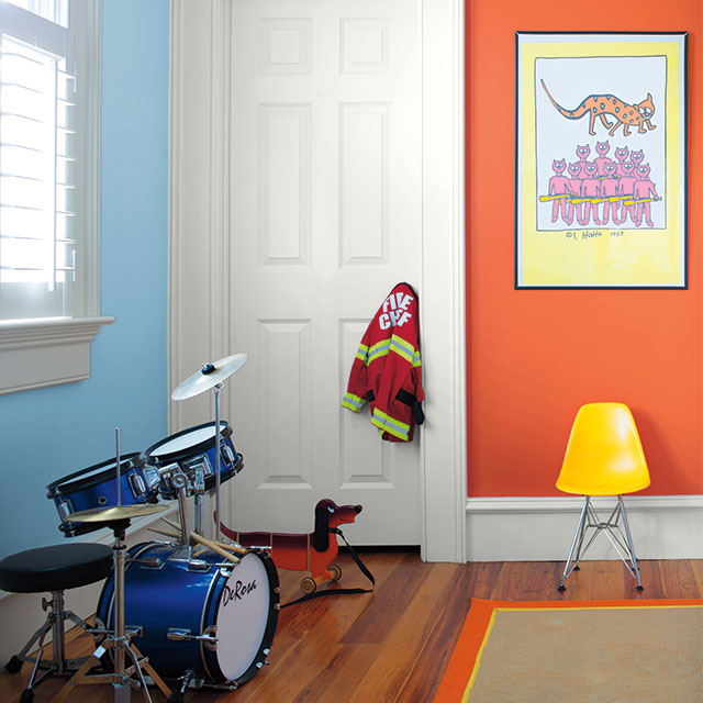 A kid’s room with one orange wall and one blue, white trim and doors, a drum set, a yellow chair, and toys.