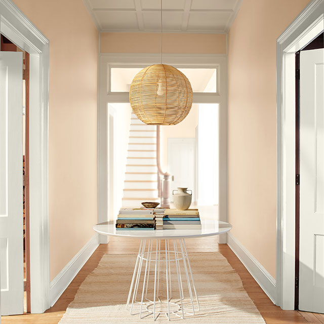 Un couloir pêche avec des portes et des moulures blanches, un luminaire suspendu en rotin, une table moderne et un tapis blanc.