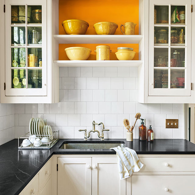 A mostly white-painted kitchen with black counter tops and orange accent behind shelving.