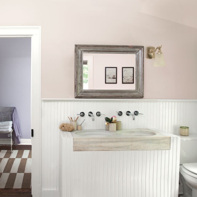 A bathroom with taupe walls tinged with light purple undertones; white wainscotting, cabinetry and trim; silver mirror; and white door open to a chair and rug in hallway.