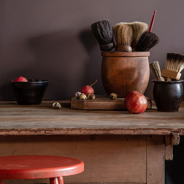 An artist’s wooden desk with pots filled with brushes, apples scattered along the tabletop, and a red stool, all in front of a purple-painted wall.