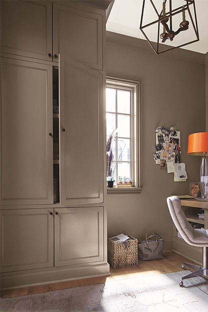 Un bureau à domicile peint en taupe avec un placard, un fauteuil beige, un bureau en bois, un babillard, un plafond blanc et une lampe abstraite.