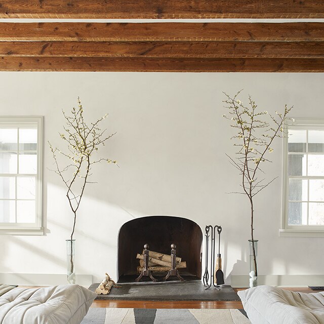 A living room painted in off-white with an arched fireplace flanked by branches in glass vases beneath a ceiling with exposed wood beams.