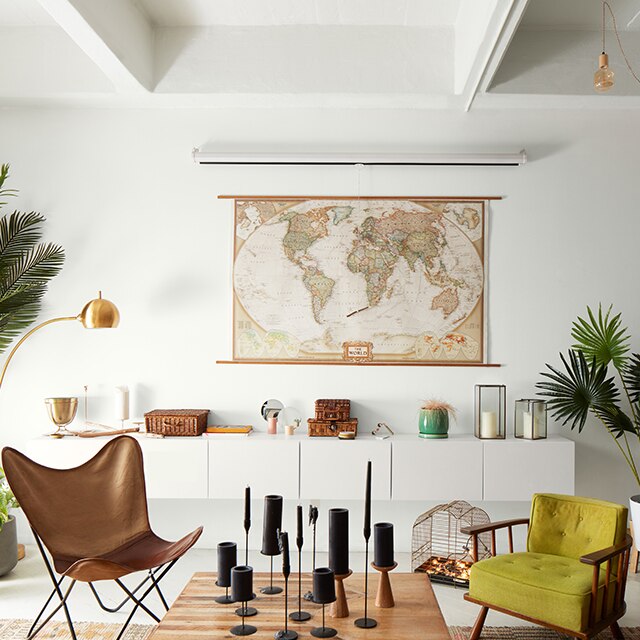 A bright loft living room painted in the same white hue using different sheens with a world map over modern wall-set cabinetry, a leather butterfly chair, a wood cocktail table with black candles, and several planters.