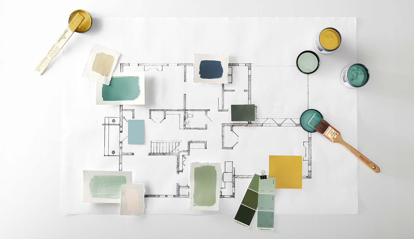 Various Benjamin Moore color samples, swatches and open jars of paint in shades of green, yellow, blue and tan, along with a paintbrush and stir stick displayed on top of a floor plan.
