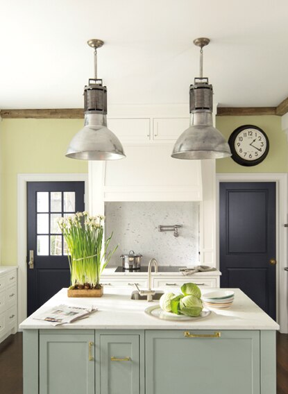 In this serene kitchen painted in soft greens and off-whites, a white, marble-topped island sits underneath two pendant lights.