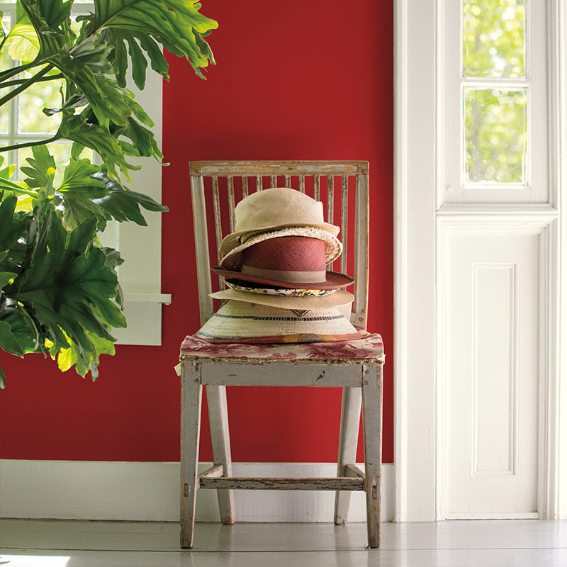 A pile of well-worn sunhats sits on a wooden chair against a red hallway wall next to an open front door.