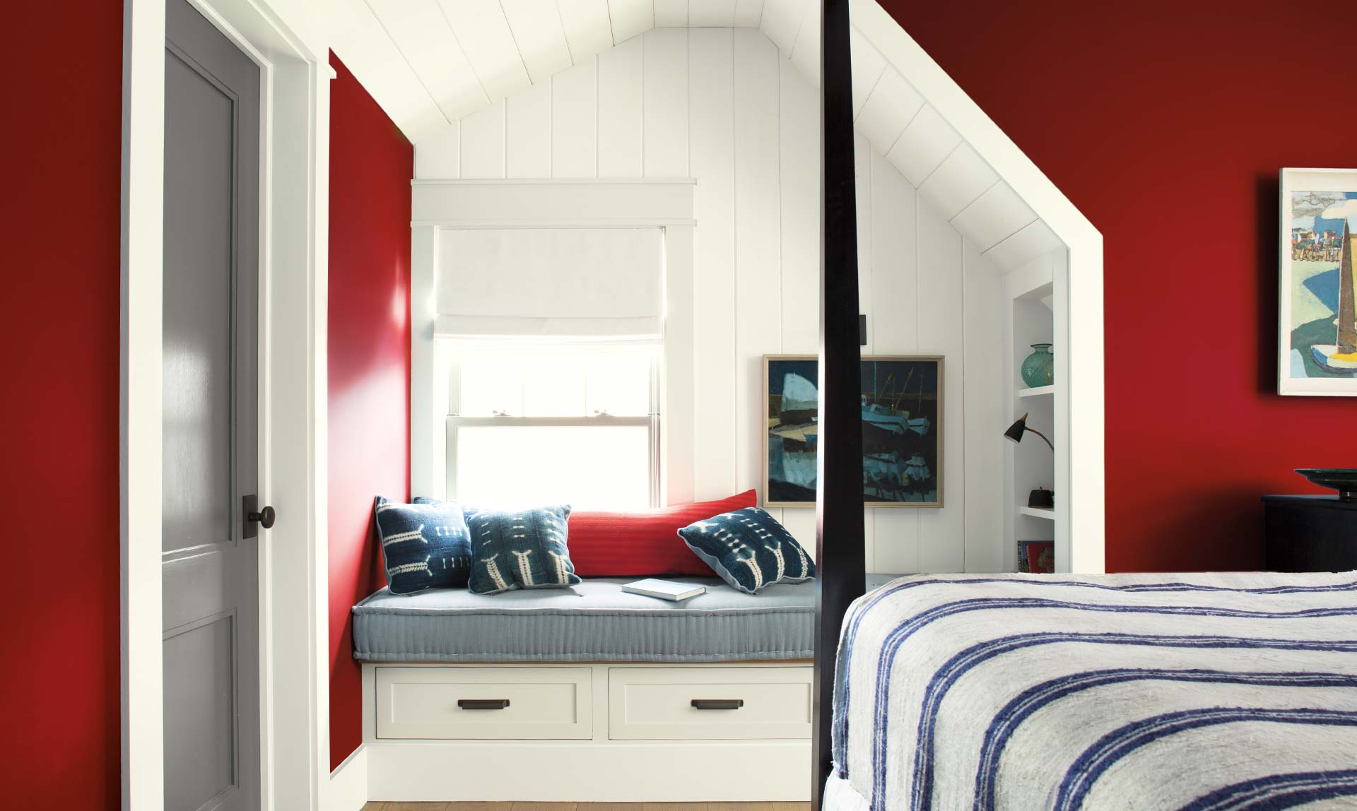 A cozy bedroom with built in cushioned bench features red walls and a poster bed.