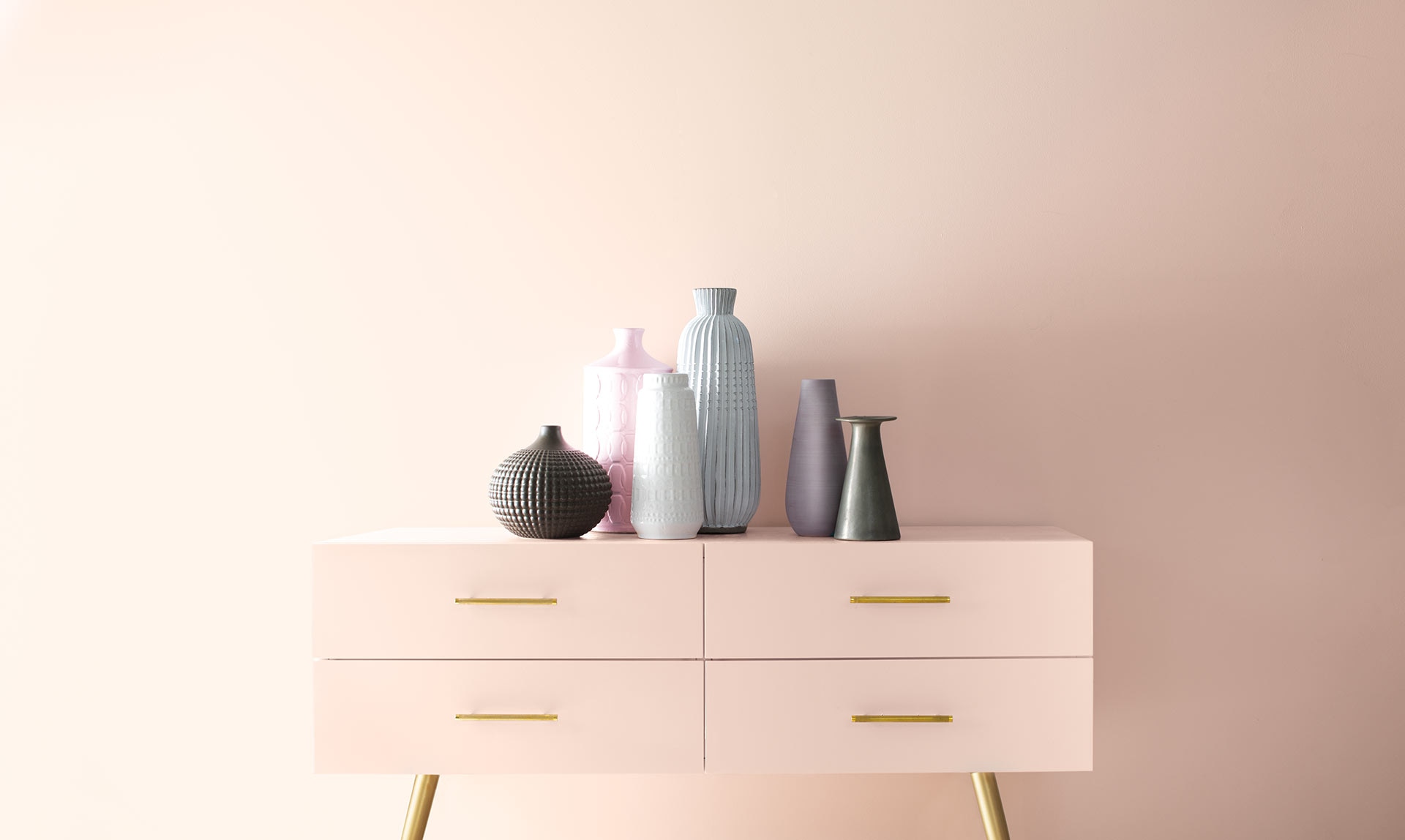 Un mur rose pâle encadre une table console rose ornée de poignées dorées où sont posés de nombreux vases