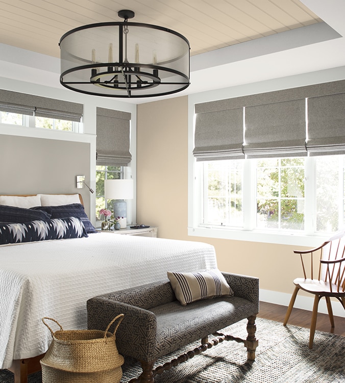 An airy bedroom with white bedding, navy and white pillows, circular chandelier, and upholstered bench.