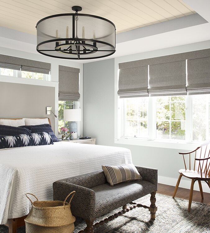 An airy bedroom with white bedding, navy and white pillows, circular chandelier, and upholstered bench.