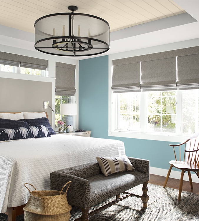 An airy bedroom with white bedding, navy and white pillows, circular chandelier, and upholstered bench.