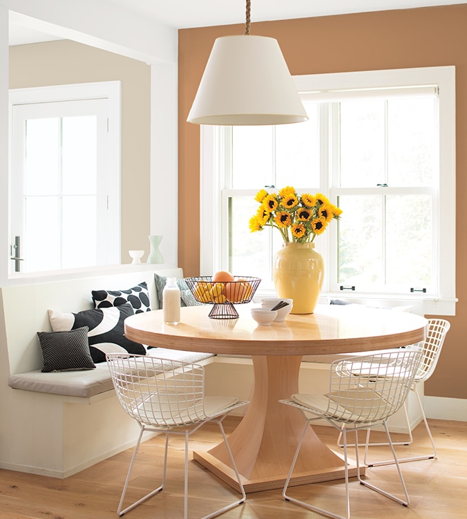 Coin-repas lumineux avec banquettes encastrées, table ronde en bois, chaises blanches en treillis et lampe suspendue blanche.