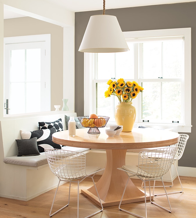 Coin-repas lumineux avec banquettes encastrées, table ronde en bois, chaises blanches en treillis et lampe suspendue blanche.